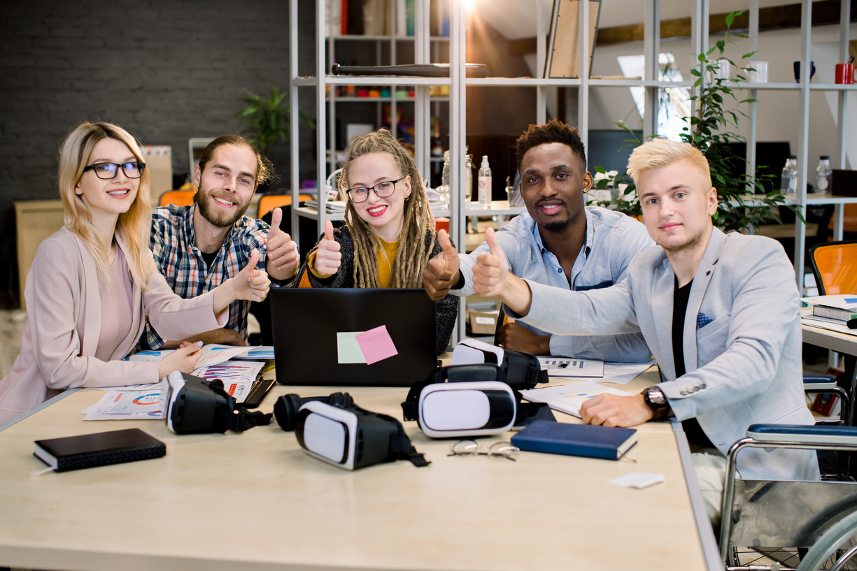 Collective work of full-fledged people and people with disabilities. Multiethnic young people sitting in office and showing thumbs up. People with disabilities in everyday life. Disabled businessman